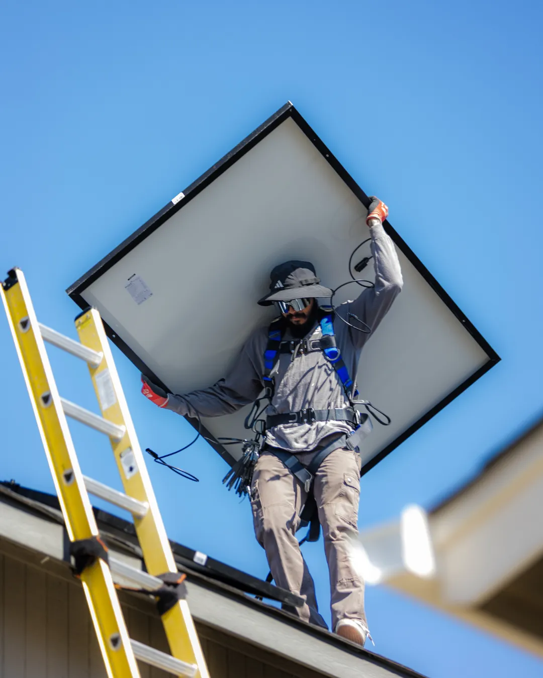 Worker on a ladder installing a large solar panel against a blue sky